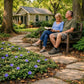 A peaceful garden scene with two people sitting on a wooden bench, one reading a book, surrounded by purple flowers, trees, and a small house in the background.