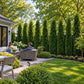 Two people sitting on a stone patio surrounded by manicured shrubs and tall evergreen trees in a well‑maintained backyard garden.

